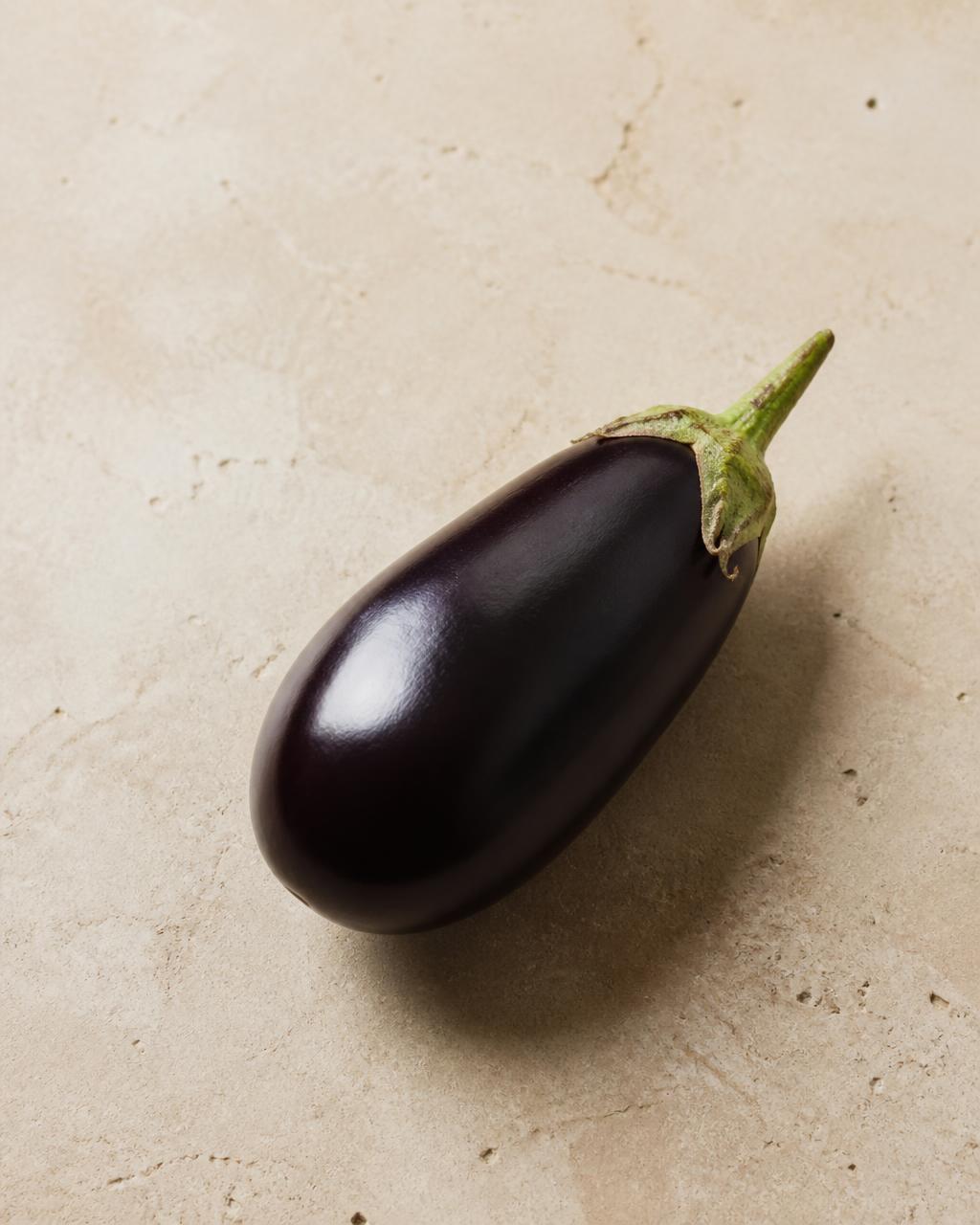 One whole glossy purple eggplant on a warm cream stone surface