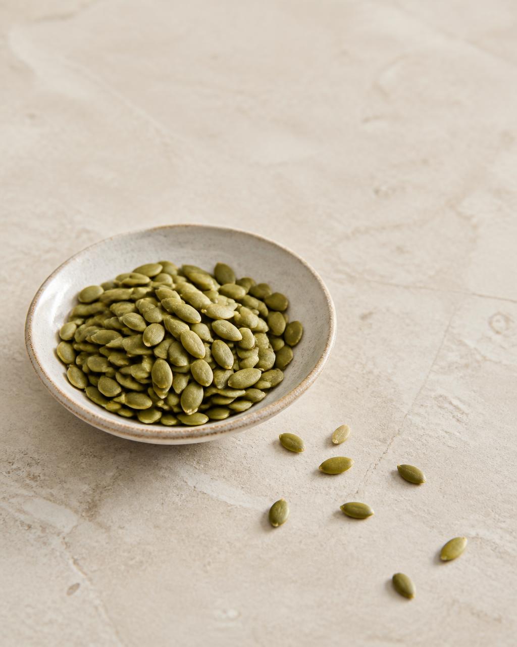 Green pumpkin seeds in a small ceramic bowl on a warm cream stone surface