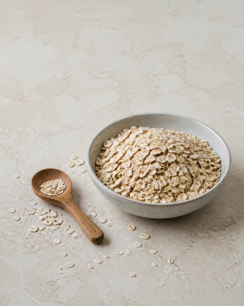 Rolled oats in a ceramic bowl with a small wooden spoon on a warm cream stone surface