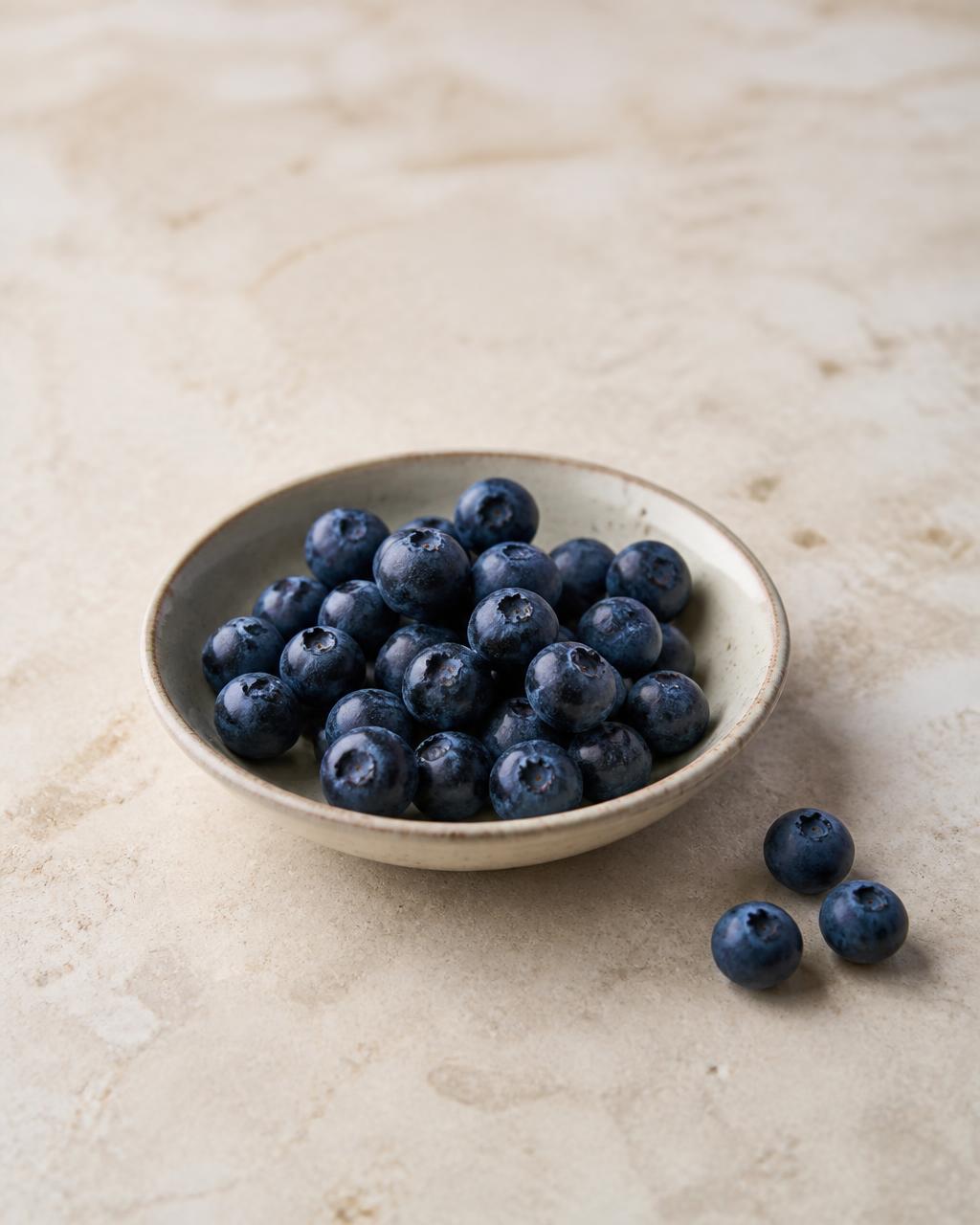 Fresh blueberries in a small ceramic bowl on a warm cream stone surface