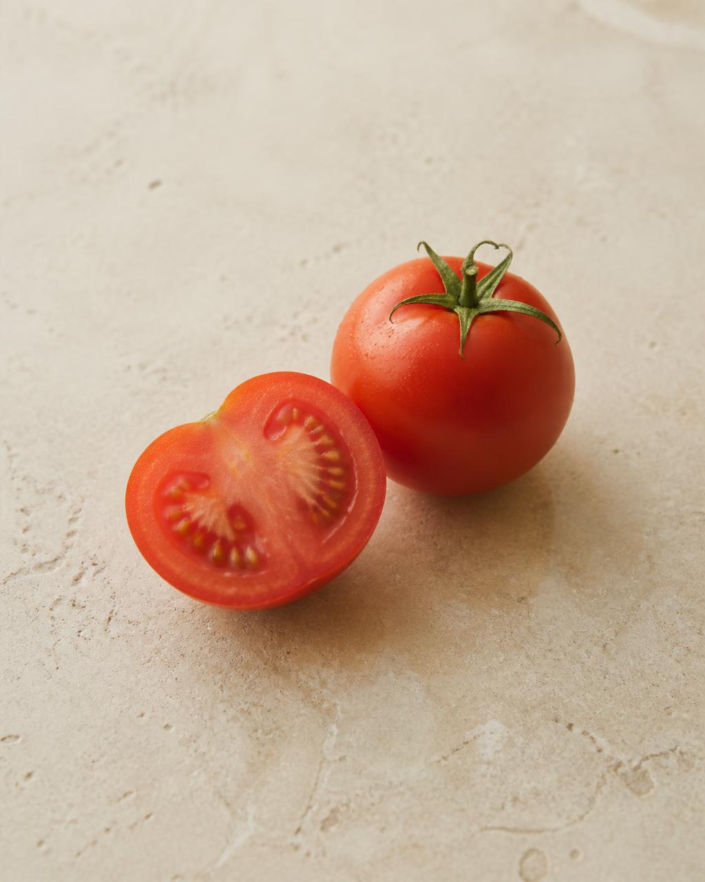 One whole and one halved ripe tomato on a warm cream stone surface
