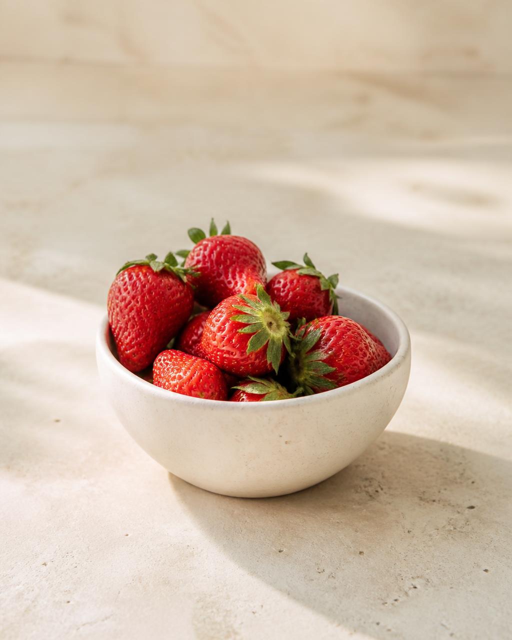 Fresh strawberries in a small pale ceramic bowl on a warm cream stone surface
